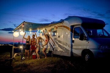 Fun with friends in camp, in front of rv. Young group of people dancing, playing guitar, and celebrating. Summertime togetherness. Travel, holiday, weekend, lifestyle concept. - 584738587