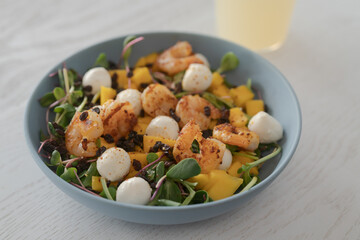 Salad with mango, mozzarella, shrimps and mixed microgreens in blue bowl on white oak table