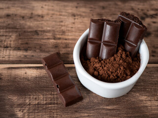 Bitter aerated chocolate bars and cocoa powder in white bowl on the wooden table. Pieces of dark porous chocolate