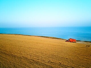 Aerial of red combine harvester working in wheat field near cliff with sea view on sunset. Harvesting machine cutting crop in farmland near ocean. Agriculture, harvesting season. Landscape scenic.