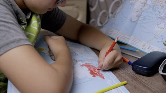 Sad Boy in glasses Drawing Country of Russia on World Map with a Red Pencil. Schoolboy is sitting at the table in bedroom, doing homework in geography. War in Ukraine. Concept of the Russian invasion.