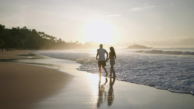 Silhouettes of a young happy couple holding hands and running on the beach together enjoying summer back view. Boyfriend and girlfriend having fun at the seaside hugging and kissing at sunrise.