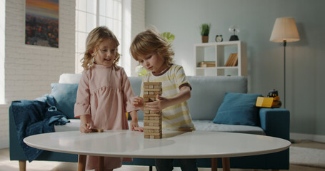 Funny cute caucasian brother and sister playing a table top game together, taking wooden bricks out of tower and applauding, happily smiling - family fun at home 4k footage