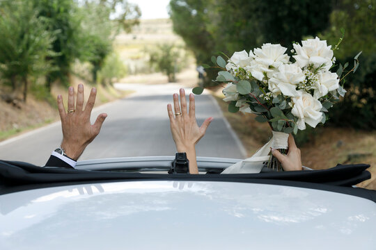 due neo sposi tirano in alto le mani ce il bouquet che esce dal tetto dell'auto in corsa