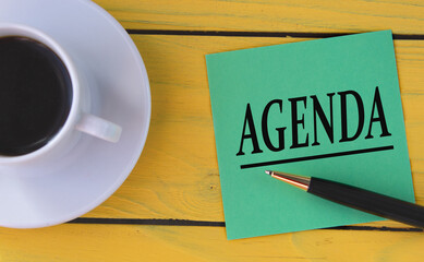 AGENDA - word on a green sheet on a yellow wooden background with a pen and a cup of coffee