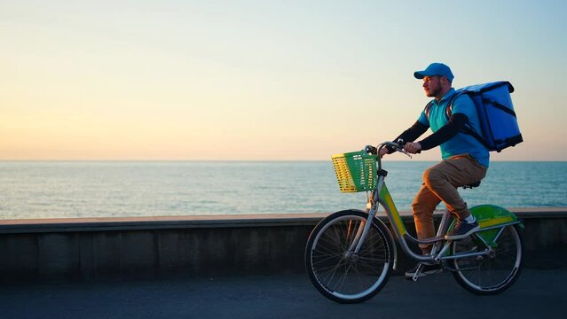 a positive deliveryman in a blue uniform with a backpack rides a bicycle , sunset or sunrise