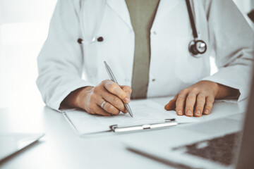 Unknown doctor woman sitting and writing notes at the desk in clinic or hospital office, close up. Medicine concept