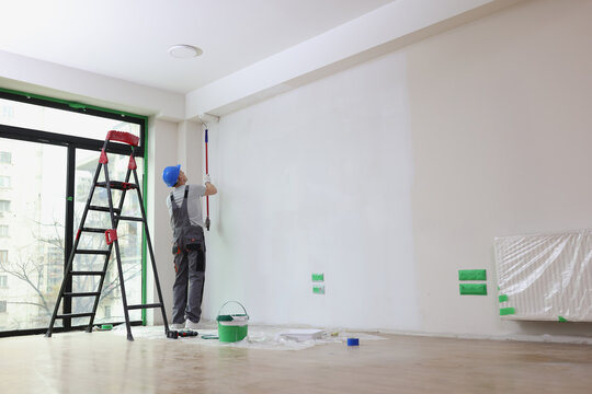 Employee with helmet paints wall with roller in premise