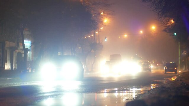 Car traffic with headlights flare on the dark night slippery street road of the city in wet cold snowy winter weather with fog in bokeh