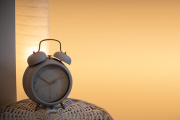 On the wicker lid of the basket stands an analog alarm clock against a background of a lit bedside lamp.