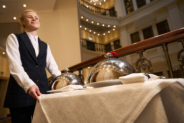 Joyful waitress carries serving cart along corridor of chic hotel