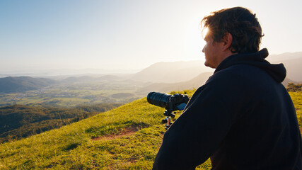 Photographer on the hill. Young male photographer takes pictures with camera and telephoto lens set on tripod on the green hill in the mountains at sunrise