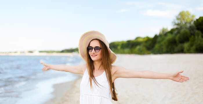 Happy smiling woman in free happiness bliss on ocean beach standing with a hat, sunglasses, and open hands. Portrait of a multicultural female model in white summer dress enjoying nature