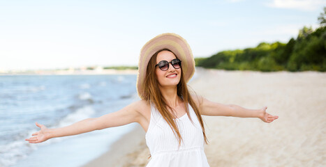 Fototapeta na wymiar Happy smiling woman in free happiness bliss on ocean beach standing with a hat, sunglasses, and open hands. Portrait of a multicultural female model in white summer dress enjoying nature