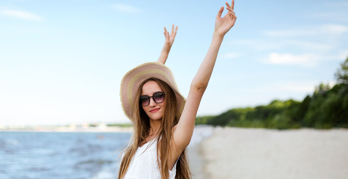 Happy smiling woman in free happiness bliss on ocean beach standing with a hat, sunglasses, and rasing hands. Portrait of a multicultural female model in white summer dress enjoying nature