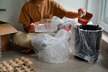 a boy of eight years old in beige clothes, sits at home in the kitchen and sorts garbage into different bags for recycling, the child separates plastic and kraton, bottles and bags