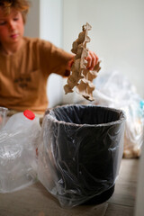 a boy of eight years old in beige clothes, sits at home in the kitchen and sorts garbage into different bags for recycling, the child separates plastic and kraton, bottles and bags