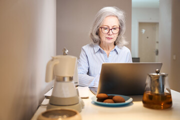 Beautiful elderly lady works with a laptop at kitchen table