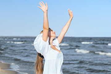 Happy smiling beautiful woman on the ocean beach standing in a white summer dress, raising hands