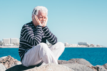 Caucasian elderly man with headphones sitting on the cliffs enjoying sea and freedom in vacation, listening music with closed eyes