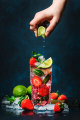 Bartender hand squeezes lime juice into strawberries mojito cocktail glass with lime, white rum, soda, cane sugar, mint, and ice in glass on deep blue background. Summer refreshing beverage