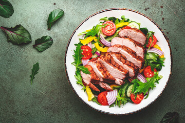 Duck salad with grilled breast and vegetables: red tomato, cucumber, paprika, onion and chard, frisse, mizuna and arugula leaves on green table background, top view