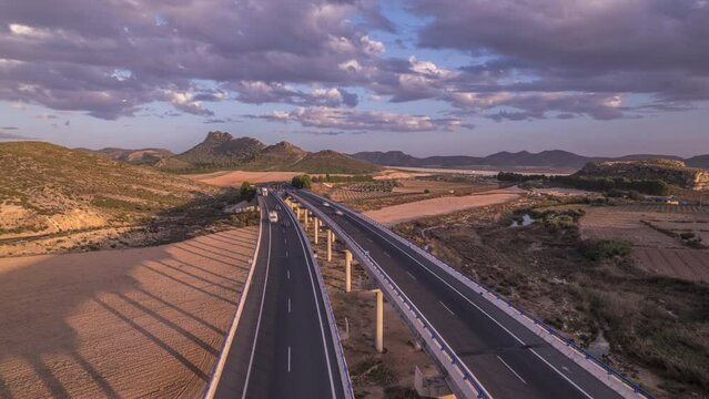 Aerial view of vehicles (car) crossing a highway in a long and high viaduct at sunset with clouds slowly change form in blue sky, Spain