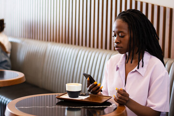 Sideways african-american woman with dreadlocks drinks coffee and checking her news feed or social media messaging using free wi-fi on mobile phone in a cafe at a table day alone in the summer
