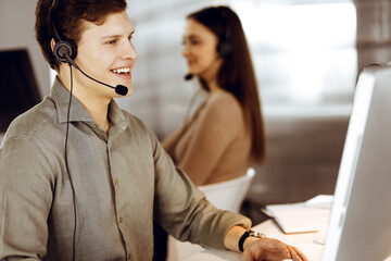 Young dark-haired guy in a green shirt and headsets is talking to a client, while sitting at the desk, working together with a female colleague in a modern office. Call center operators at work