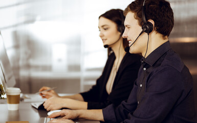 Obraz premium Two young people in headsets are talking to the clients, while sitting at the desk in a modern office. Focus on man in a blue shirt. Call center operators at work