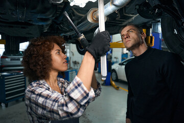 Obraz na płótnie Canvas Young woman auto mechanic and client inspect car from below