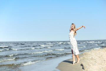 Happy blonde beautiful woman on the ocean beach standing in a white summer dress, open arms