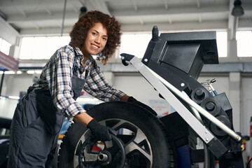 Naklejka na ściany i meble Smiling young woman with curly hair checking wheel in car workshop