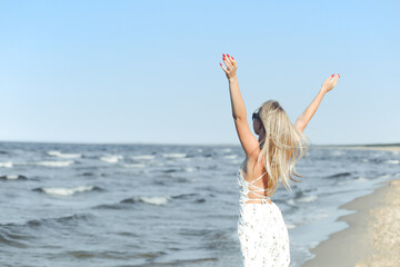 Happy blonde beautiful woman on the ocean beach standing in a white summer dress and glasses, raising hands