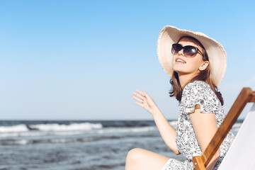 Fototapeta na wymiar Happy brunette woman wearing sunglasses and hat relaxing on a wooden deck chair at the ocean beach