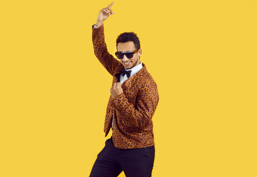 Funny African American man wearing a leopard party suit, bowtie and trendy sunglasses dancing in the studio. Portrait of a happy young black guy dancing isolated on a yellow colour background