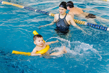 Preschool boy learning to swim in pool with foam noodle with young trainer