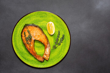 Baked salmon steak with herbs and lemon on bright round plate on black stone table top view.Healthy tasty food concept.