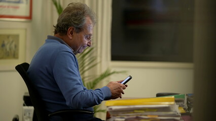 Fototapeta premium Candid mature man looking at smartphone device at home office. One older perosn in 70s working using cellphone reading message
