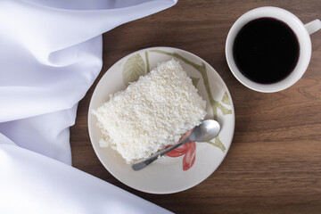 PEdaço de cuscuz em pires de porcelana sobre mesa de madeira, prato típico brasileiro, feito com tapioca, leite, açucar e leite de coco. Com copo de café and white towel.