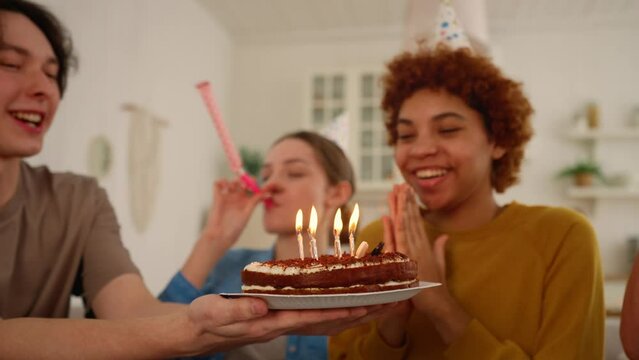 African american girl celebrates birthday with friends blows candles on cake makes a wish. They smile and clap hands. Wearing festive caps and blows party horns. Celebration party for birthday girl.