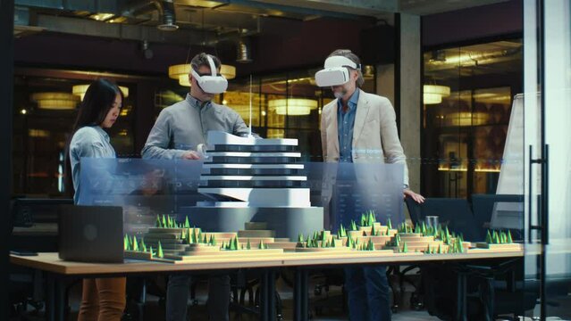 Male architect uses VR headset and wireless controllers, presents modern hi-tech building and park architectural project in virtual reality to colleagues. 3D hologram. Future digital AI technologies.