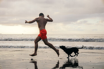 Obraz premium Young man playing with a dog on a beautiful beach at sunset