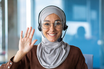 Closeup photo portrait of young beautiful Muslim business woman, tech support worker in hijab and glasses, customer service employee smiling and looking at camera, using headset , video call.