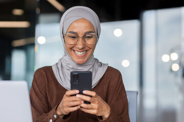 Businesswoman in hijab at work inside modern office close-up, Muslim woman using mobile phone, typing message and browsing social media at workplace, woman happy closeup