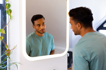Biracial man looking at himself in bathroom mirror - obrazy, fototapety, plakaty