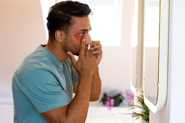 Biracial man looking in mirror and applying under eye mask in bathroom