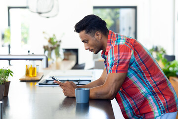 Happy biracial man leaning on countertop and using smartphone in kitchen