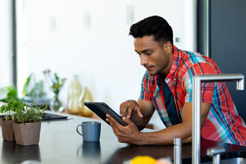 Happy biracial man leaning on countertop, having coffee and using tablet in kitchen