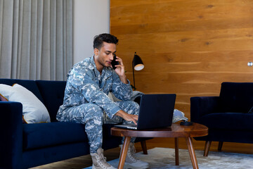 Biracial male soldier wearing uniform, using laptop and talking on smartphone in living room